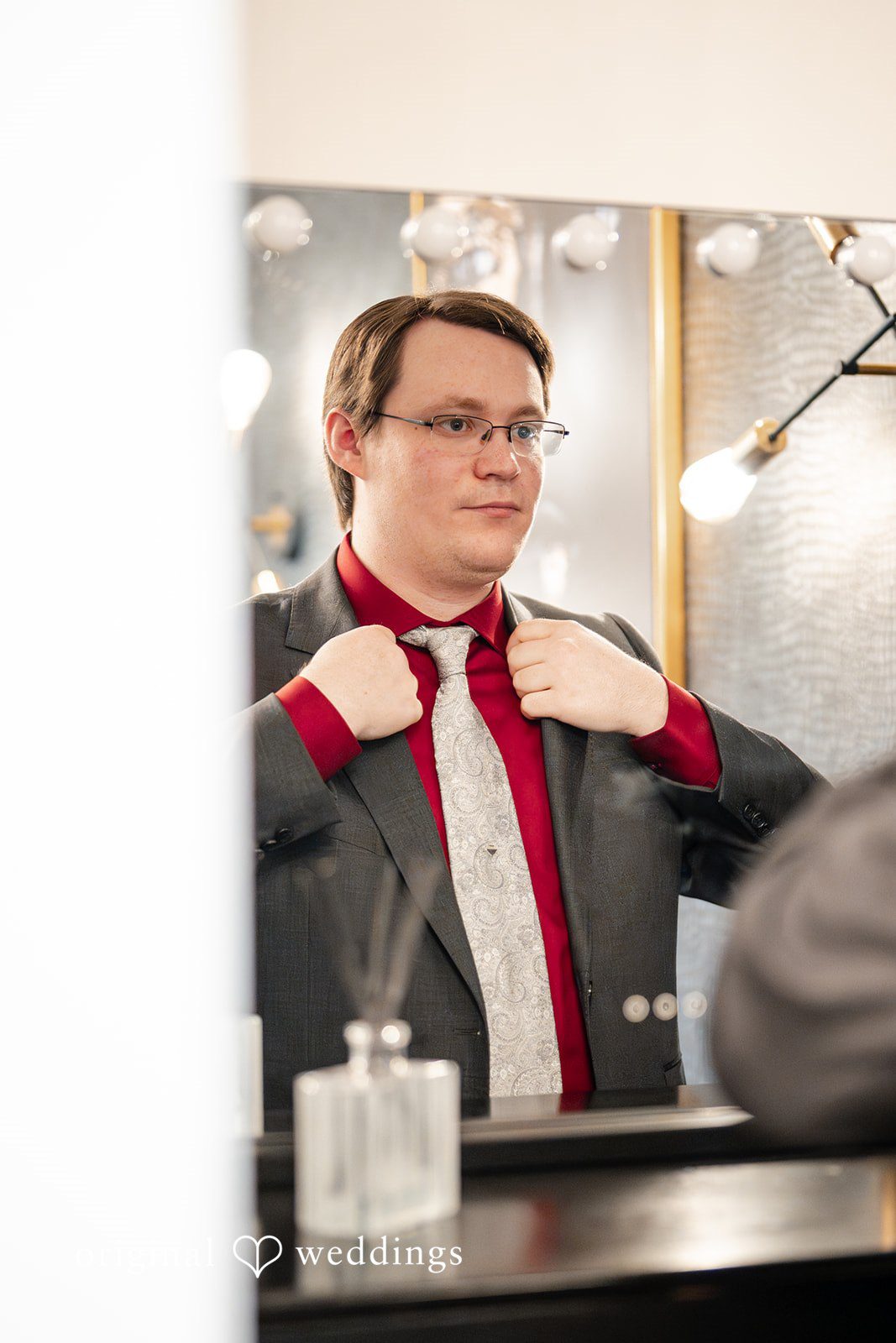 Anthony + Simi A groom in a suit adjusting his tie in front of a mirror.