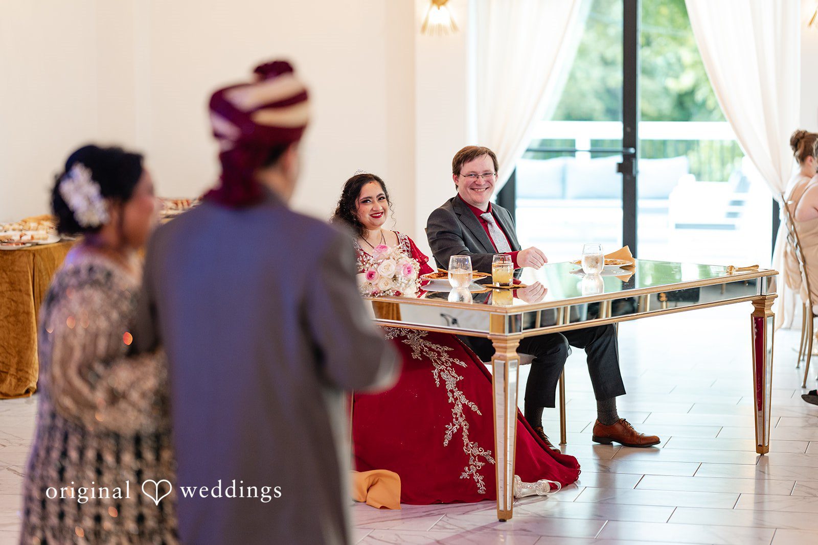 Anthony + Simi The newlyweds sit at a mirrored head table smiling as guests approach them during their reception.