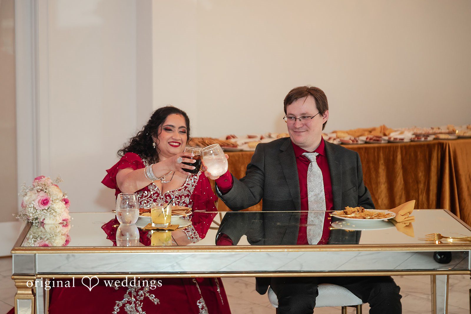 Anthony + Simi A man and a woman smiling while sitting at a reception table.