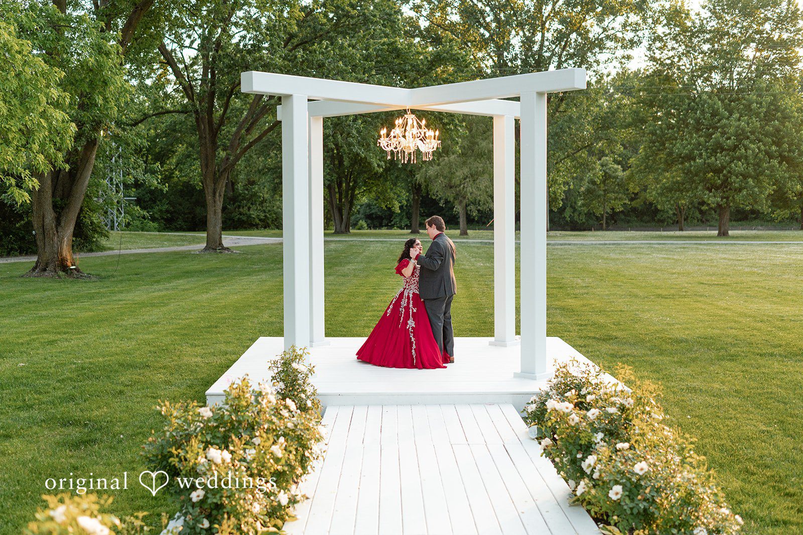 Anthony + Simi A woman in a red gown standing under a white wooden pergola with a man.