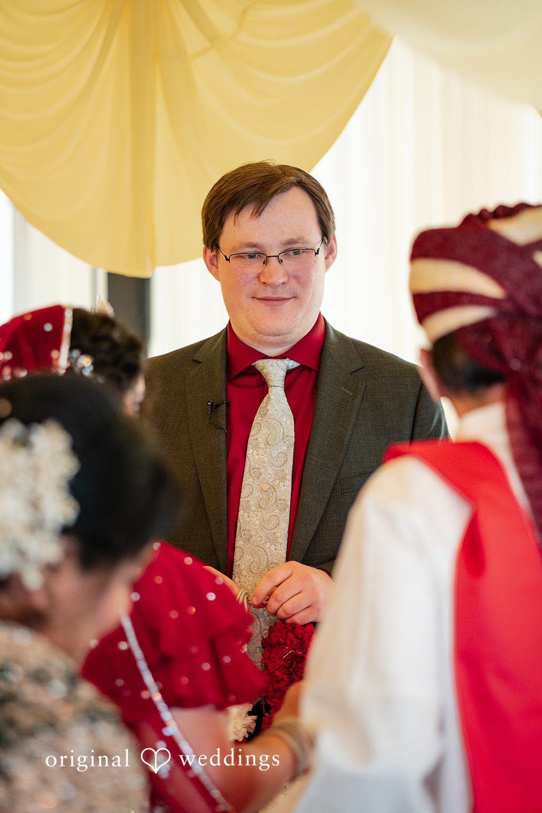 Anthony + Simi The groom stands at the altar wearing a dark charcoal suit jacket, a bright crimson button-down shirt, and a light-patterned champagne tie.