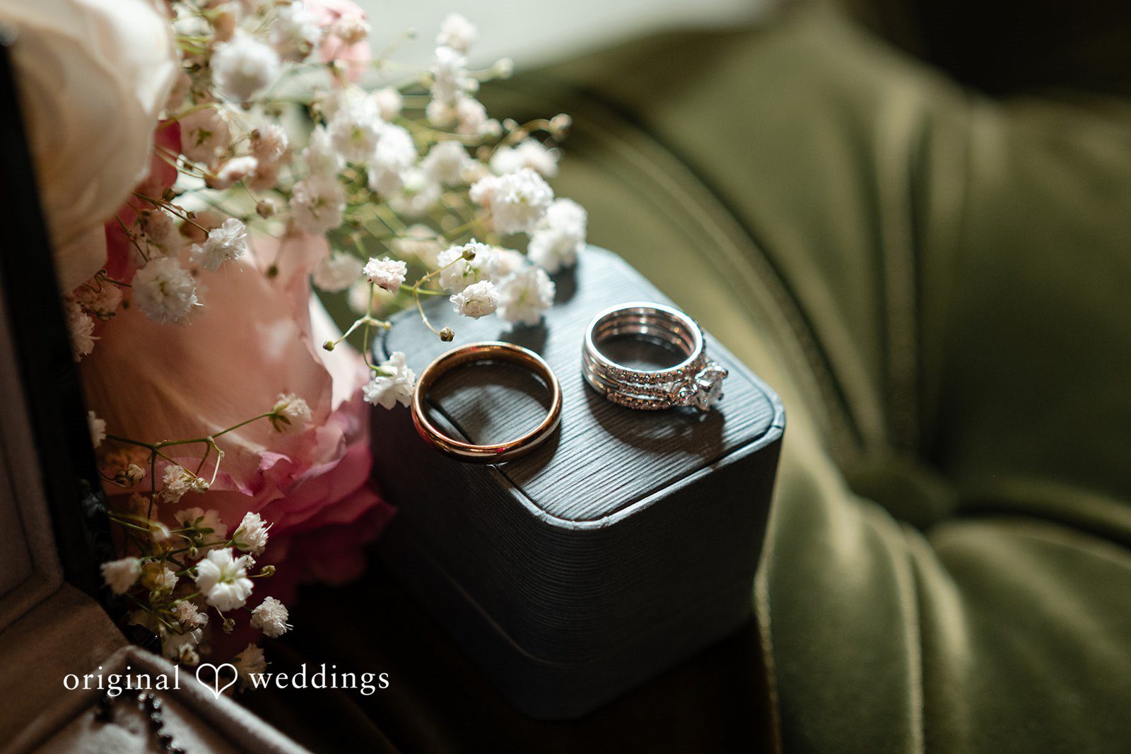 Anthony + Simi A pair of wedding rings rests on a dark textured ring box next to a bouquet of soft pink roses.