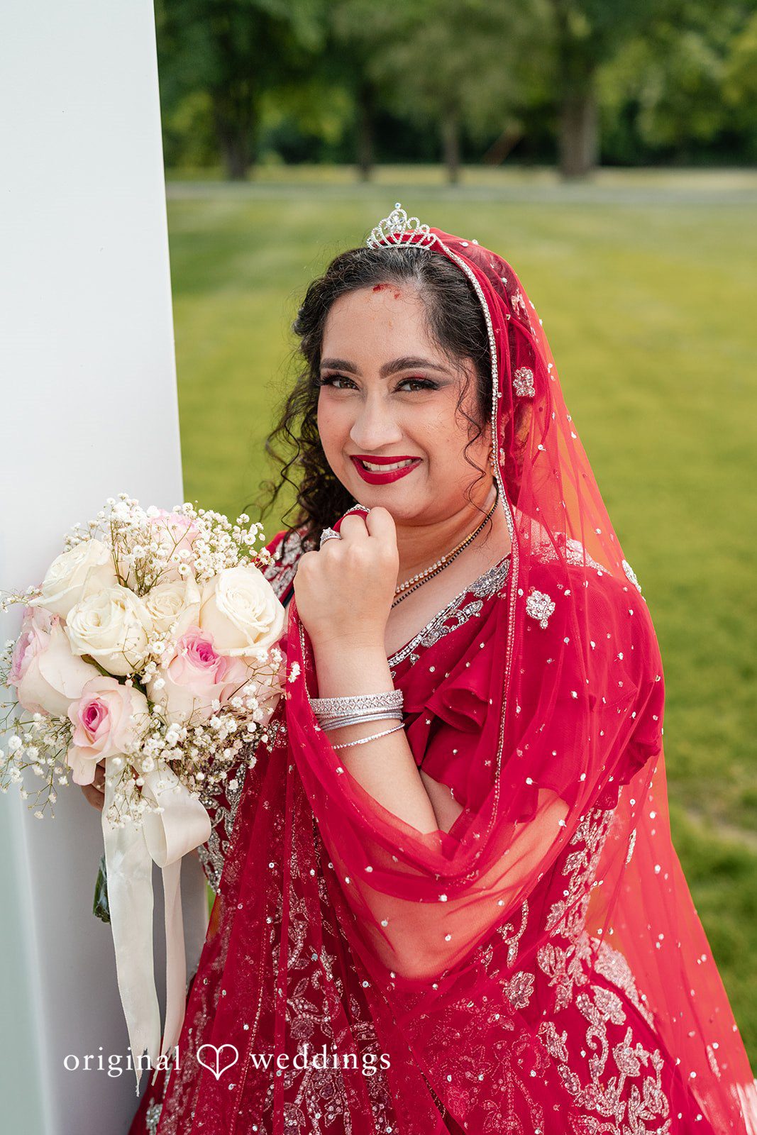 Anthony + Simi A smiling bride wearing a red outfit and holding a bouquet.