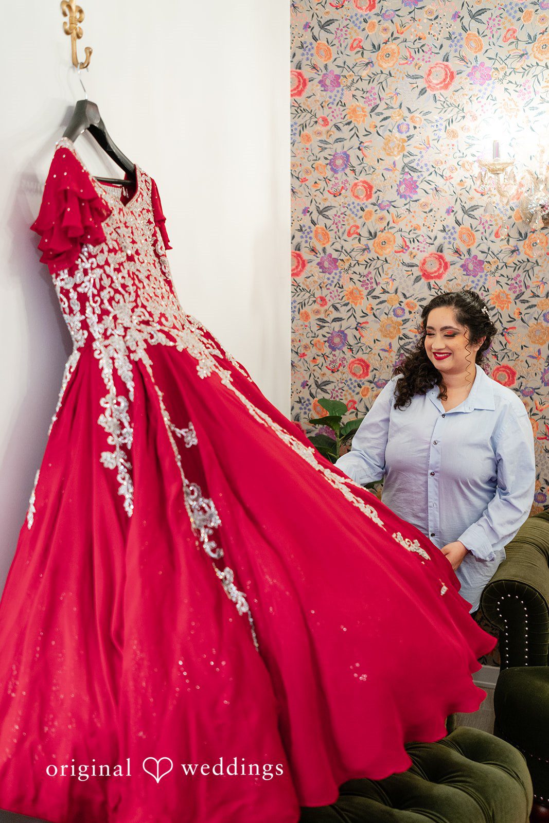 Anthony + Simi An elegant red bridal gown hangs on a wall while a woman admires it.