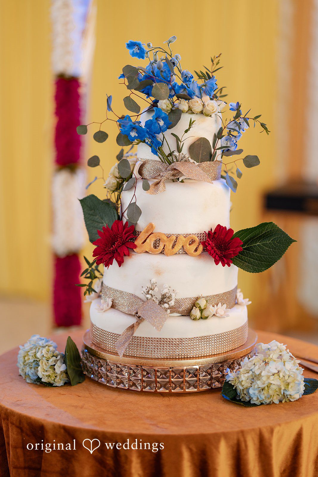 Anthony + Simi A three-tiered white wedding cake decorated with blue and red flowers.