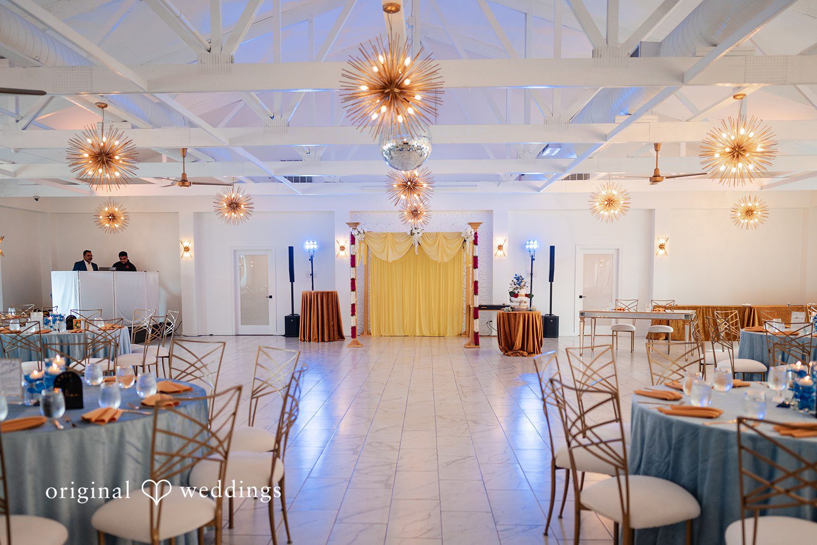 Anthony + Simi An empty wedding reception hall with tables arranged along the sides.