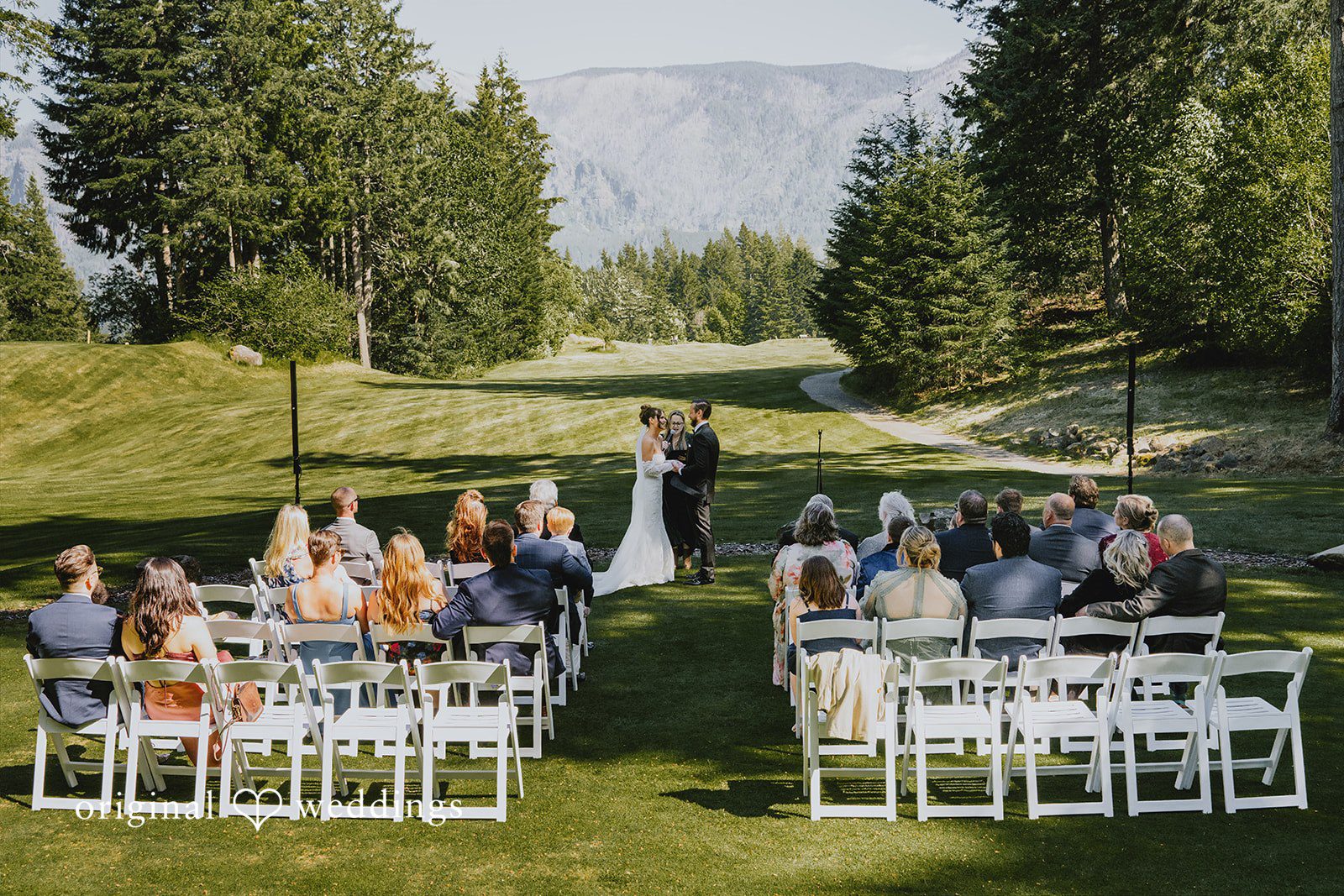 Kristina + Christopher Wide shot of an wedding ceremony with guests seated in white chairs on a lawn.