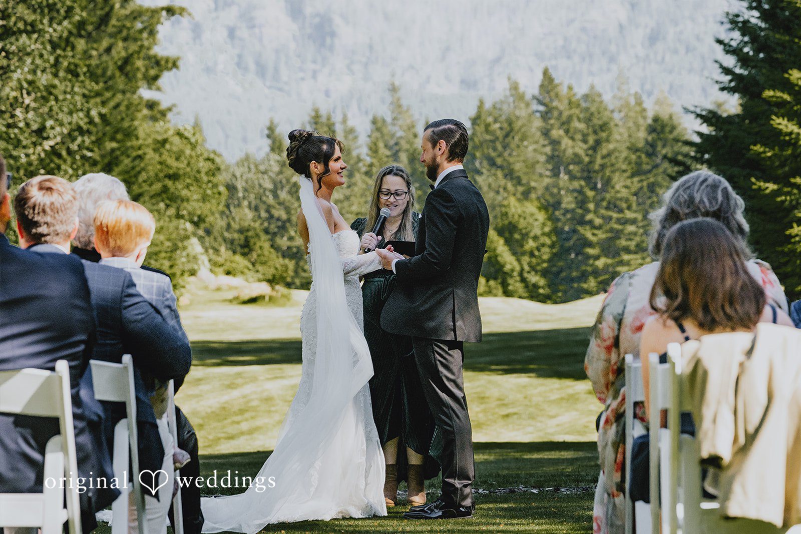 Kristina + Christopher Bride and groom holding hands during their wedding ceremony in front of trees.