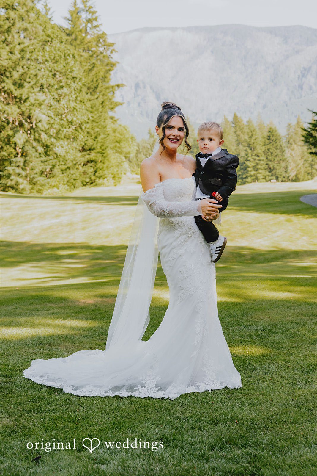 Kristina + Christopher A bride in her wedding dress with a child in a grassy field.