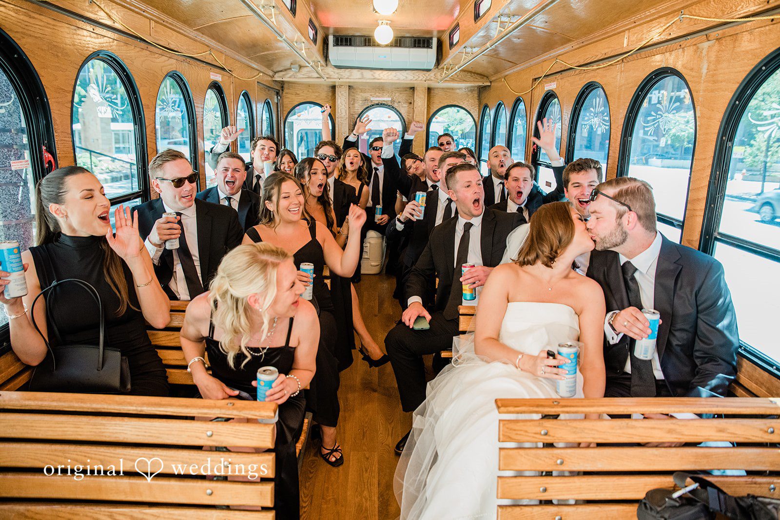 Luke + Madison Bride and groom sharing a kiss while friends celebrate inside the wedding trolley.