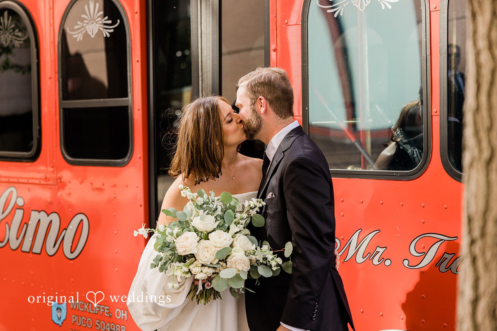 Luke + Madison Newlyweds sharing a kiss beside a bright red wedding trolley.