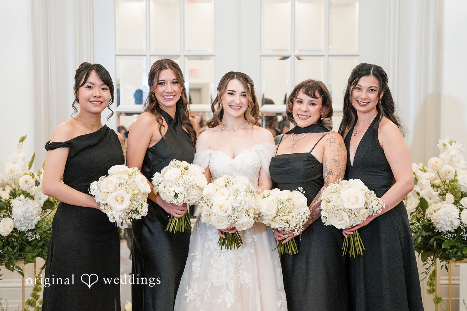 Bride posing with bridesmaids dressed in elegant black gowns and holding white bouquets.