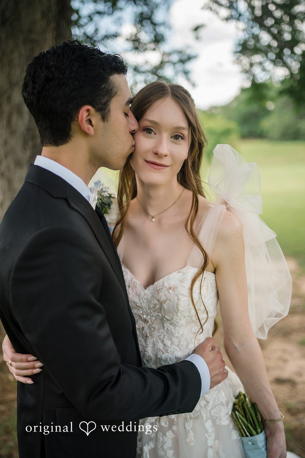 Morgan + Luke A groom in a black suit kisses the cheek of his bride in an embroidered gown with tulle shoulder bows.