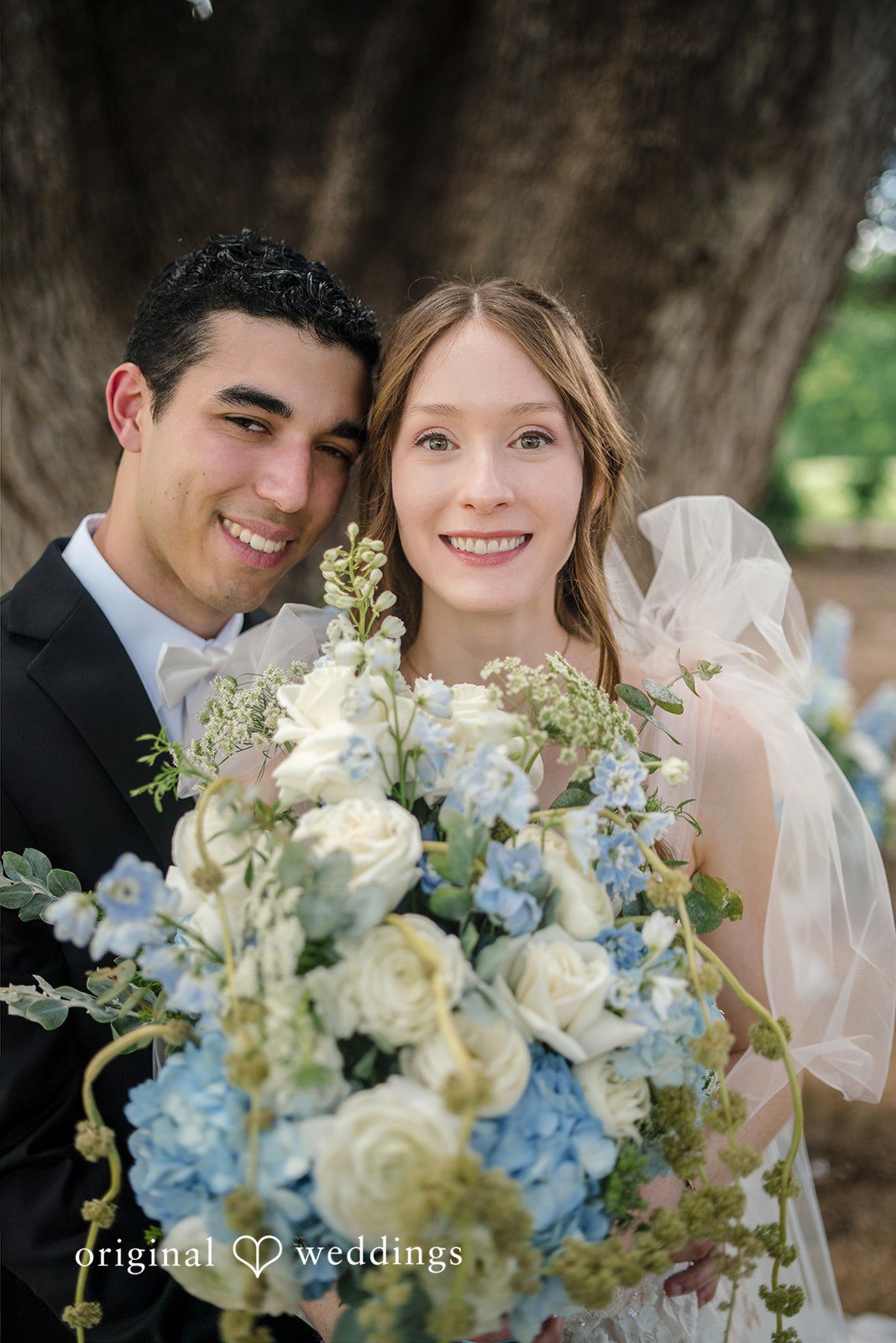 Morgan + Luke A smiling bride and groom pose closely behind a large floral arrangement.