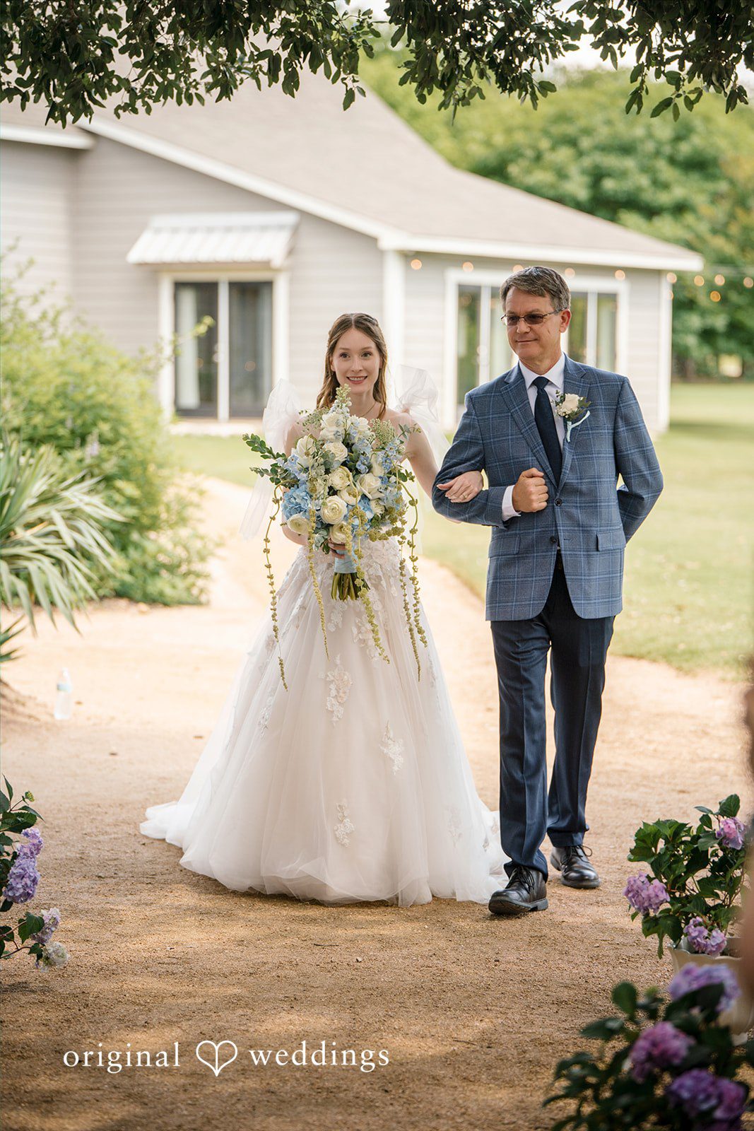 Morgan + Luke A bride walks down a paved pathway accompanied by a man at Stonebridge Wedding & Event Venue.