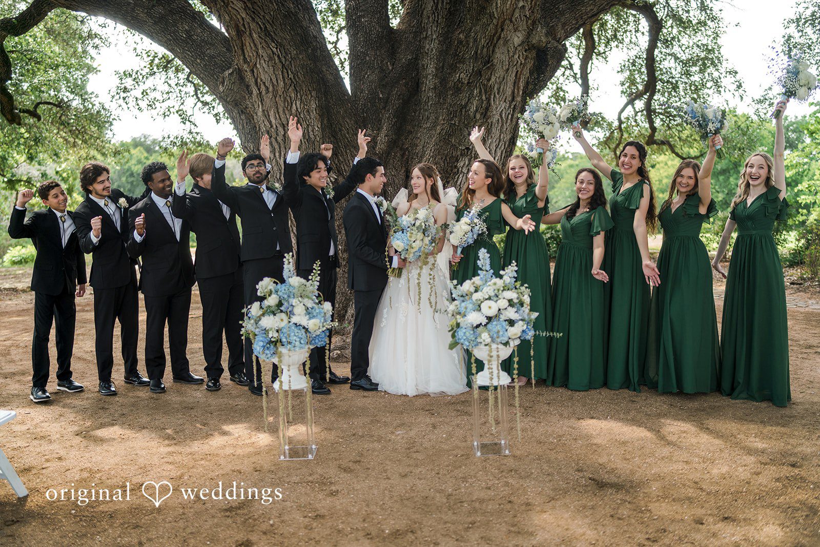 Morgan + Luke A wedding party poses together under a large tree with central blue flowers.
