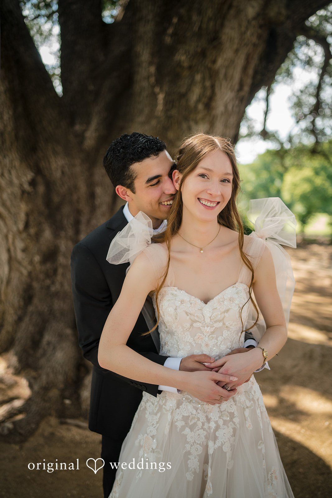 Morgan + Luke A smiling bride in an embroidered gown stands in front of a large tree while her groom embraces her from behind.