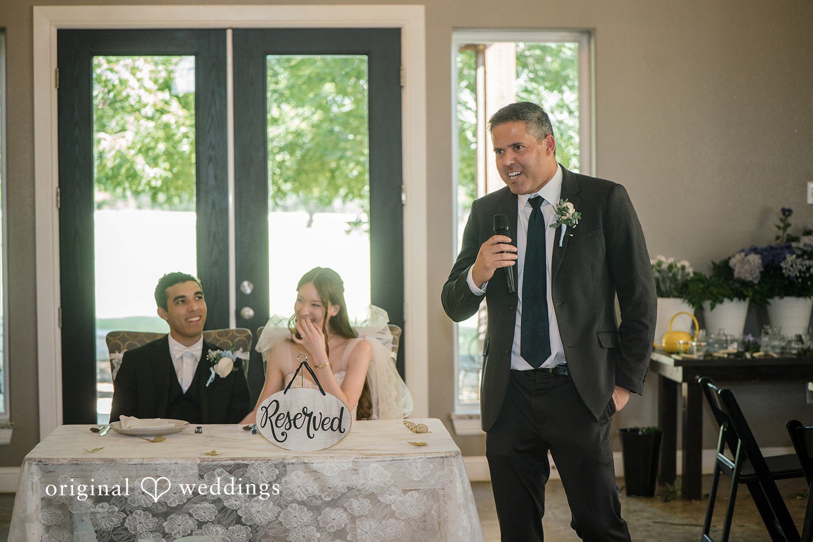 Morgan + Luke A man speaks at a microphone next to a seated couple during a reception.