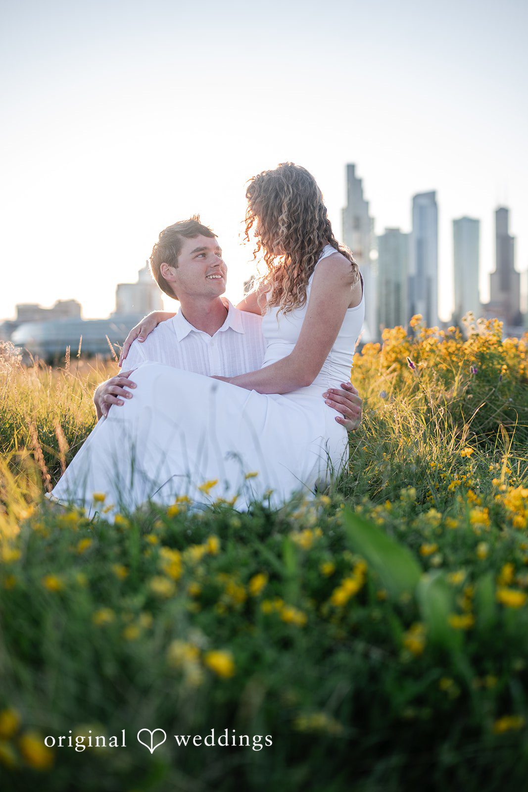 A man and a woman sitting on a grassy slope with a distant city skyline.