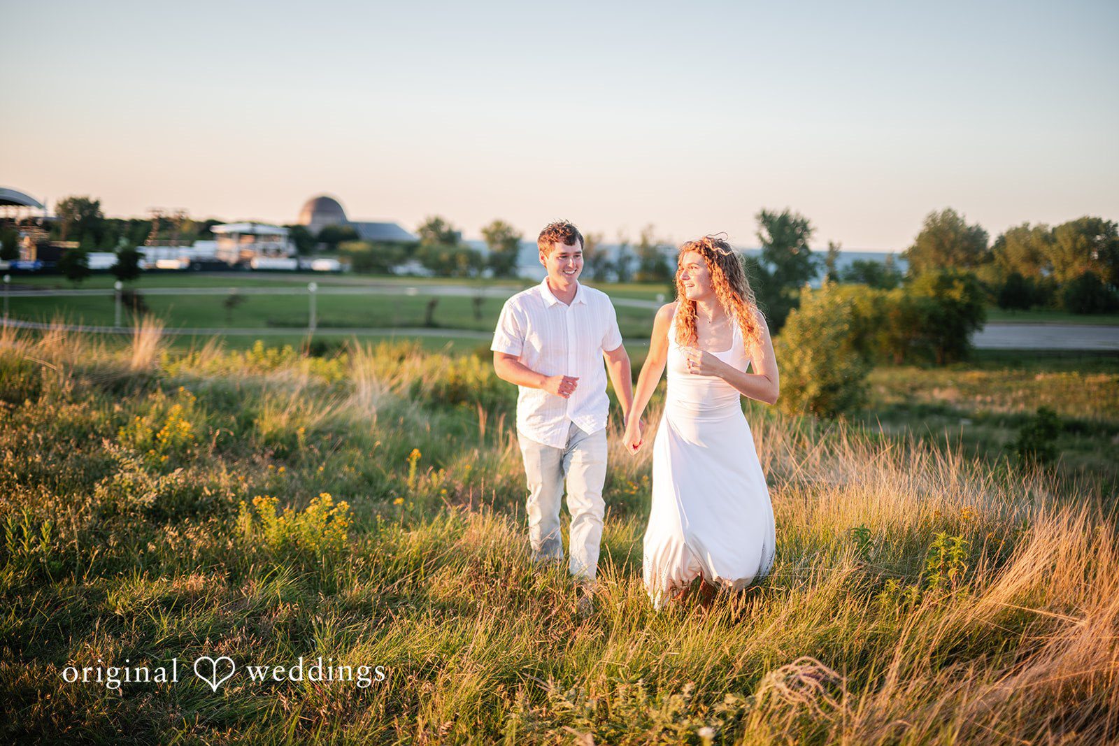 Couple in white attire walking side-by-side in a grassy landscape.