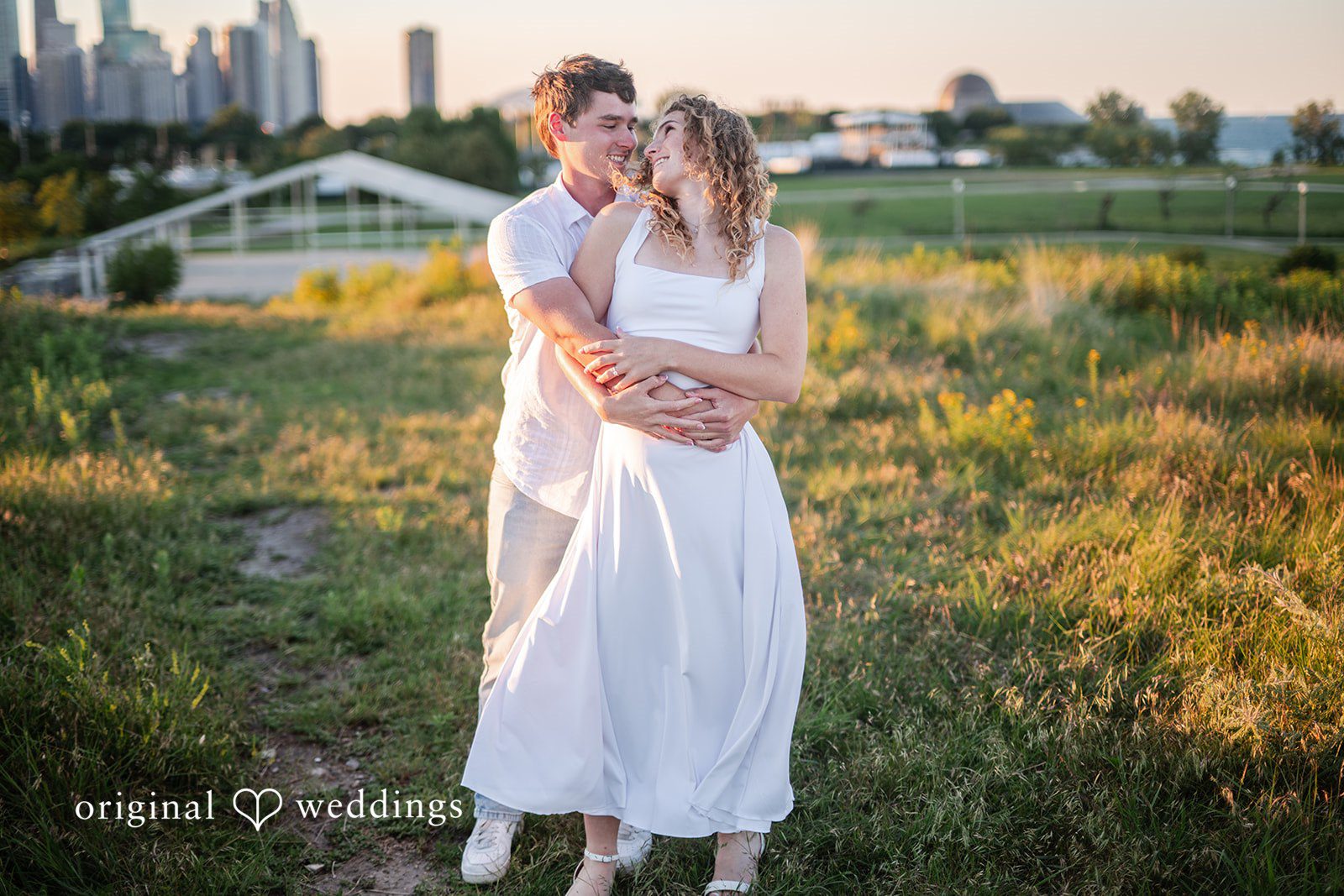 A man standing closely behind a woman dressed in a white gown in a field.