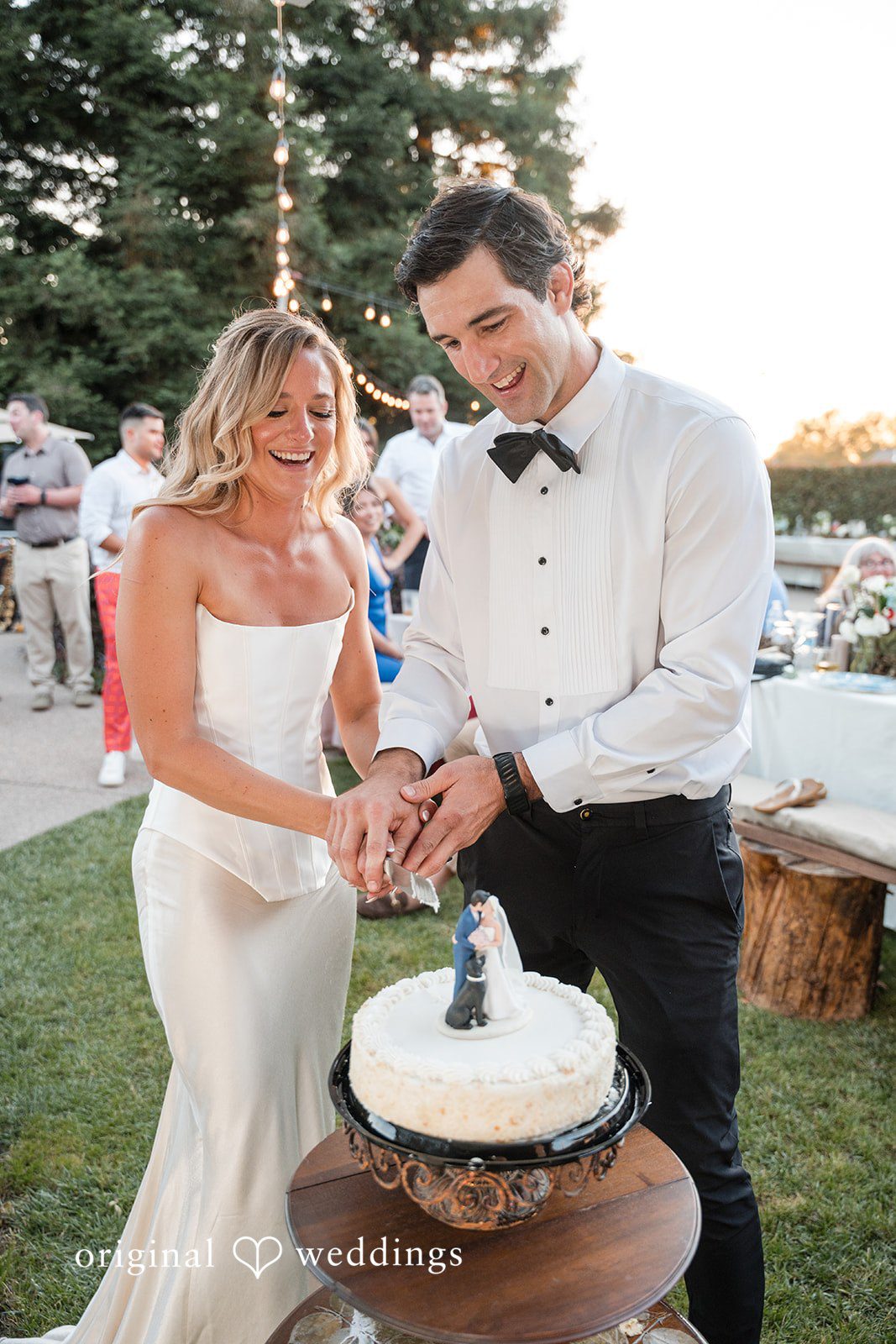 Avery + Ryan A bride and groom laughing together while slicing into their multi-tiered white wedding cake.