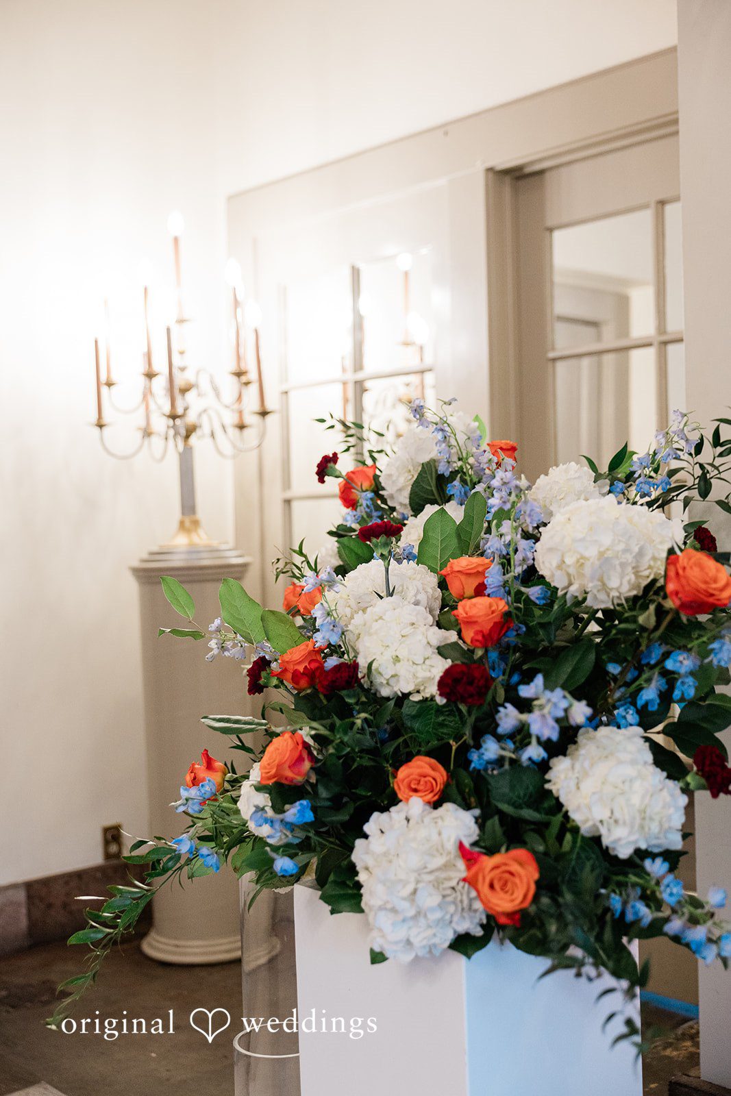 Madison + Malik A large, colorful floral arrangement next to a tall, classic candelabra.