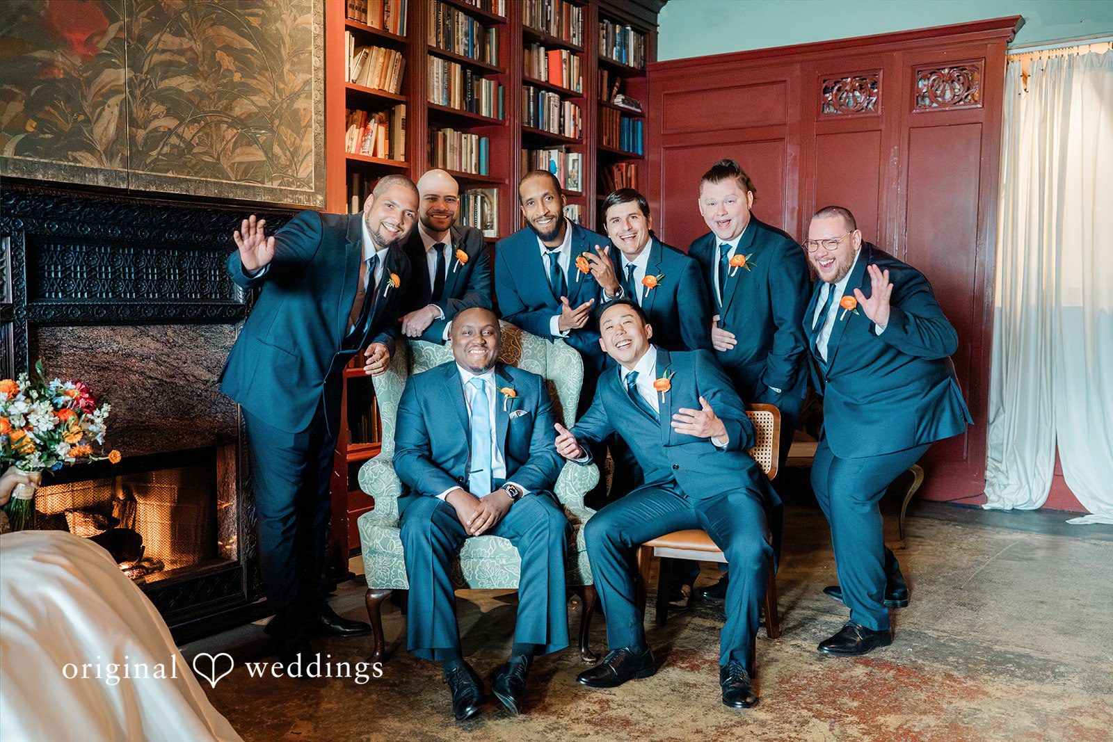 Madison + Malik Groom and groomsmen in dark suits and orange boutonnières pose playfully by a fireplace and a large bookcase.