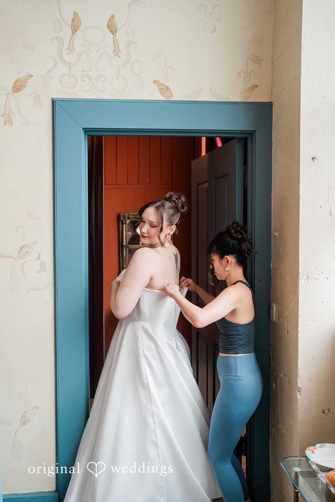 Madison + Malik A women assisting a bride with the back of her white wedding dress in a doorway.