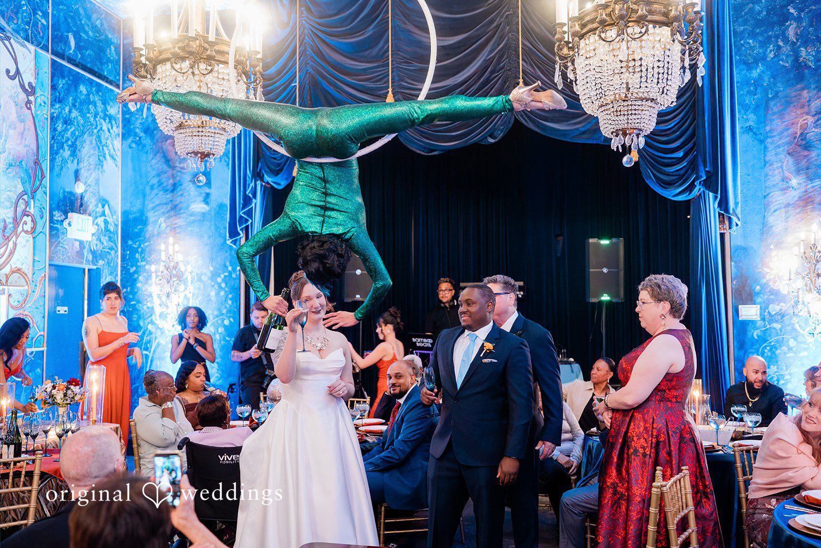 Madison + Malik An aerialist hanging upside down from a chandelier over a crowded reception hall.