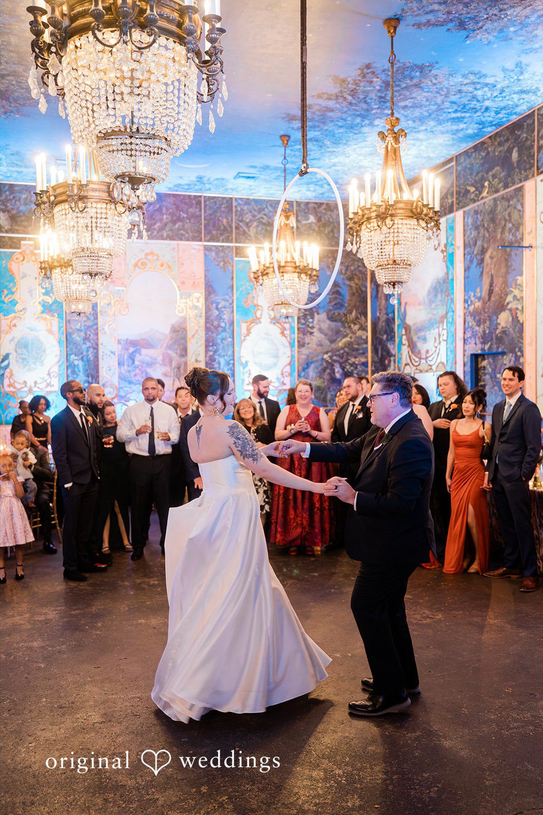 Madison + Malik A bride and a man share a dance on a dance floor surrounded by wedding guests under large crystal chandeliers.
