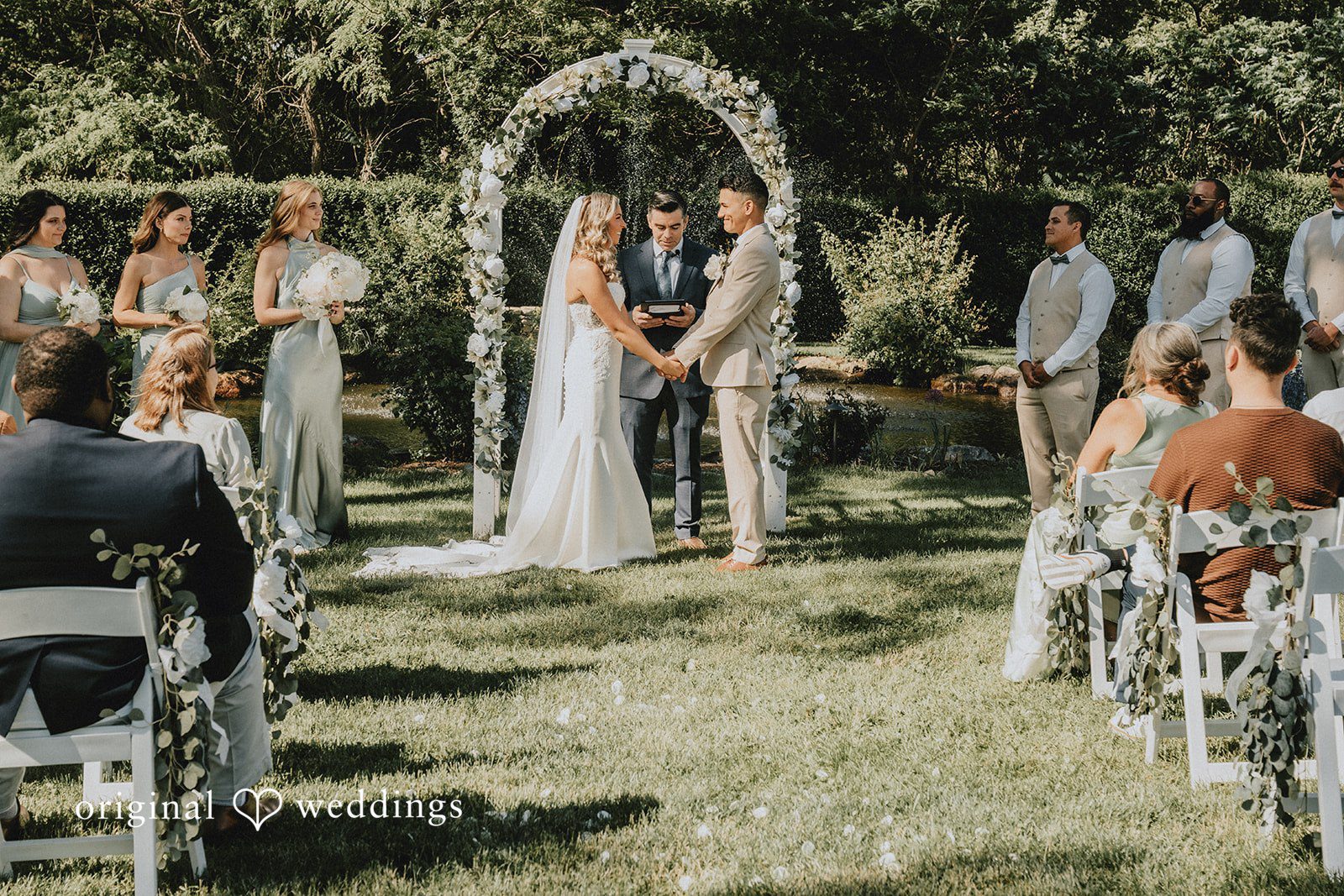 Kaylee + Raul A bride and groom standing beneath a floral arch during their ceremony while guests watch.