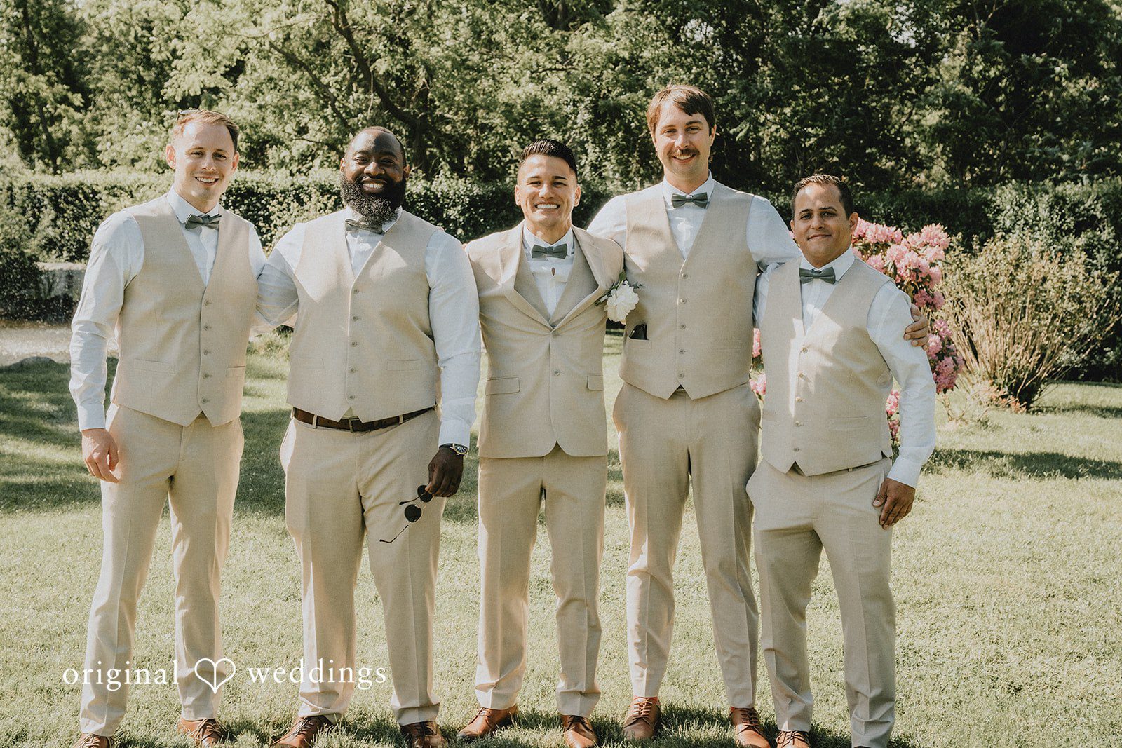 Kaylee + Raul Groom and groomsmen in matching tan vests and trousers standing in a row on a lawn.