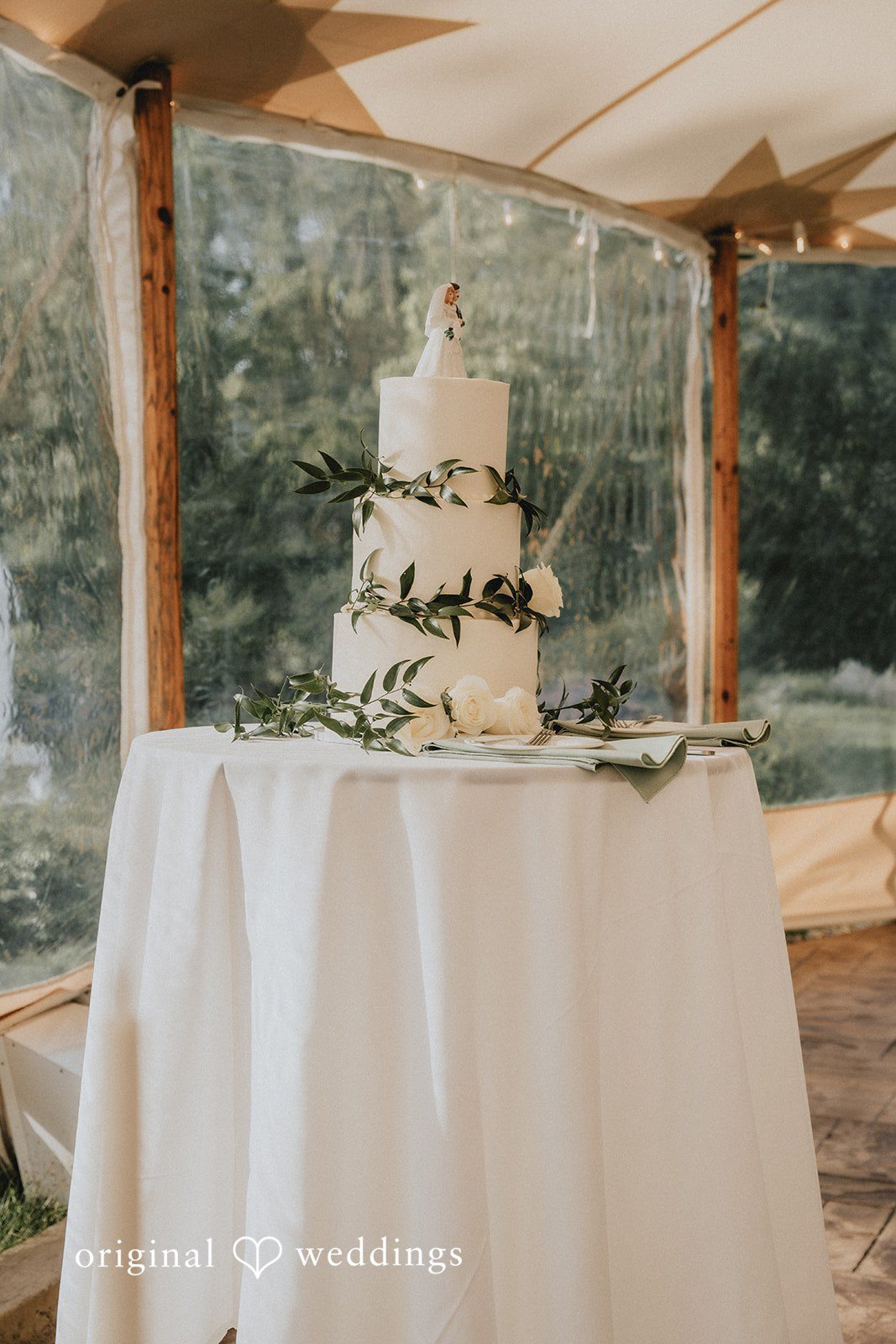 Kaylee + Raul A multi-tiered white wedding cake displayed on a table under a rustic wooden structure.