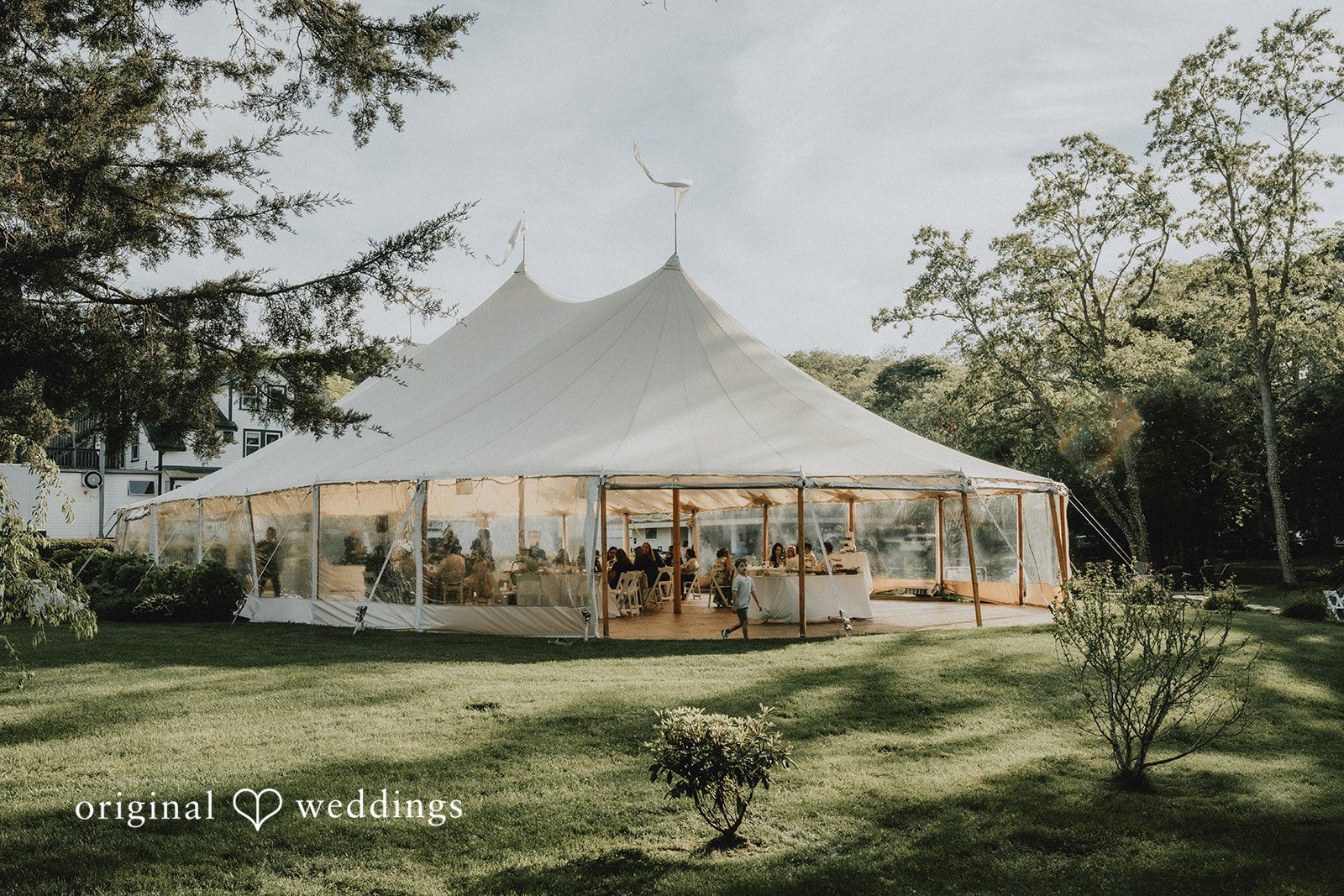Kaylee + Raul A large white wedding tent set up on a grassy lawn surrounded by trees.
