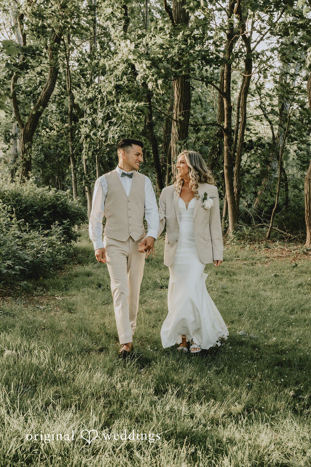 Kaylee + Raul A bride and groom walking hand-in-hand along a grassy path surrounded by trees.