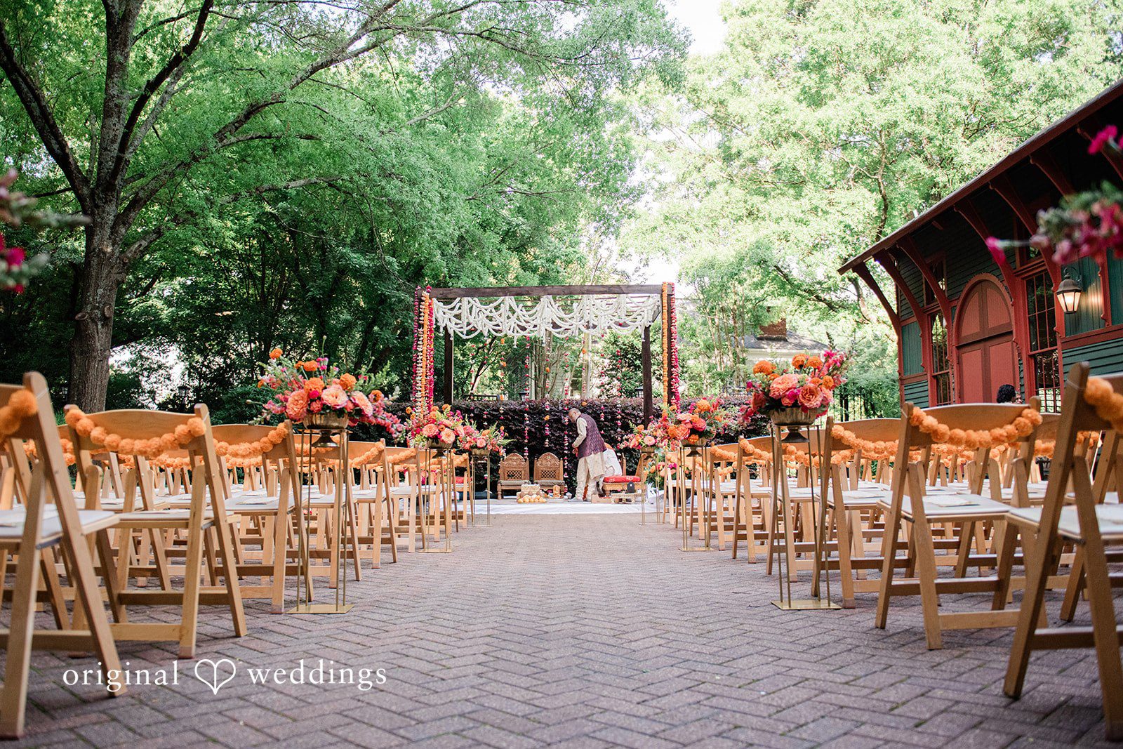Sonya + Akshaya A wedding aisle lined with chairs leading to a floral mandap among trees.