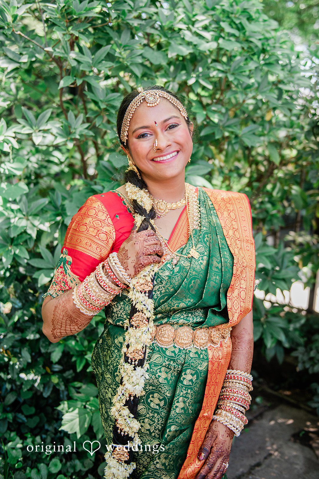 Sonya + Akshaya A bride smiling warmly while posing surrounded by lush green foliage.
