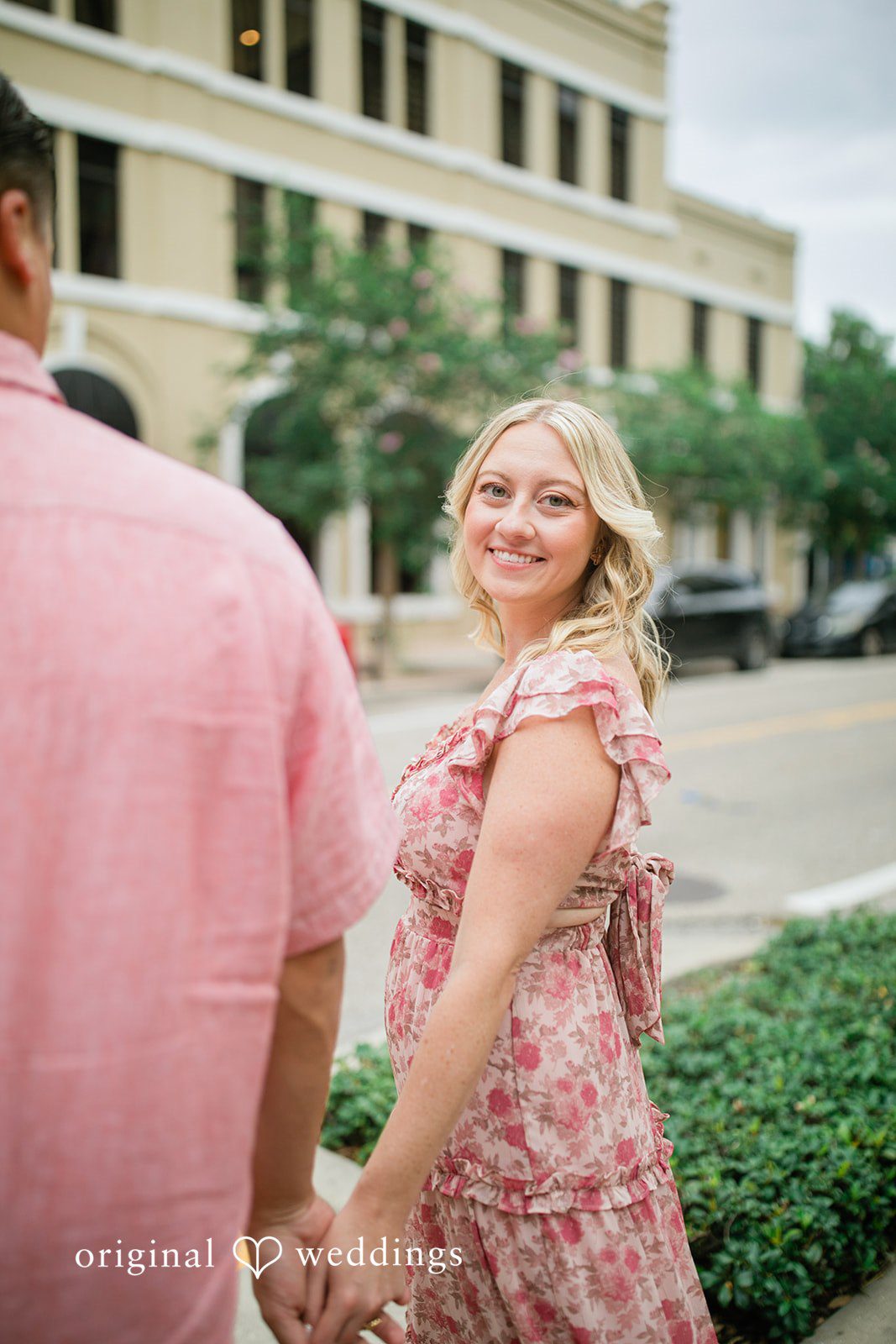 Taylor + Jack A couple walking hand-in-hand, with the woman in a pink floral dress looking back toward the camera.
