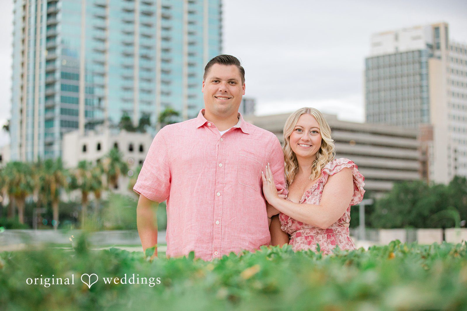 Taylor + Jack Couple posing behind green bushes with a city skyline behind them.