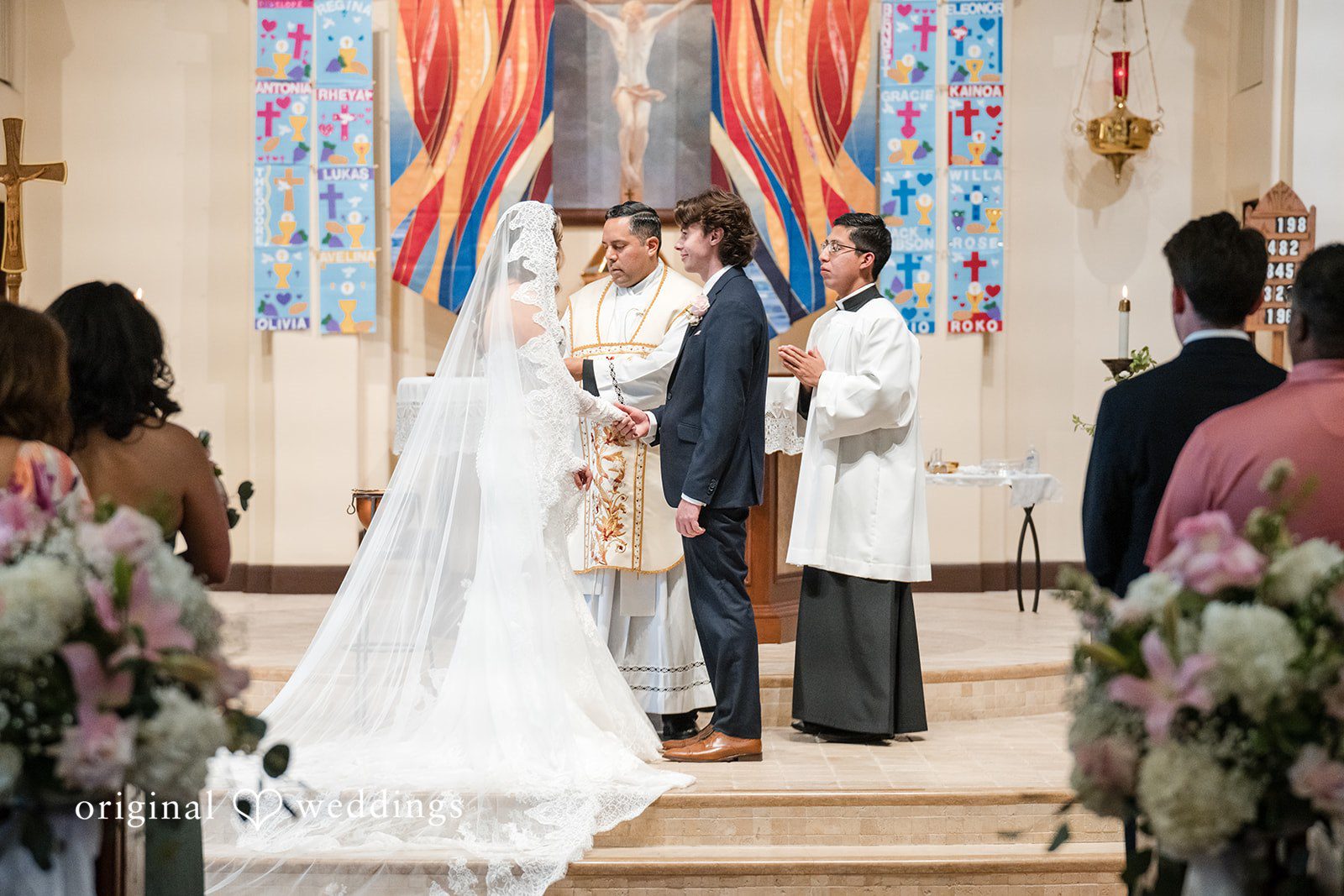 Maya + Max A side view of a bride and groom exchanging vows in front of a priest and wedding guests at the altar.