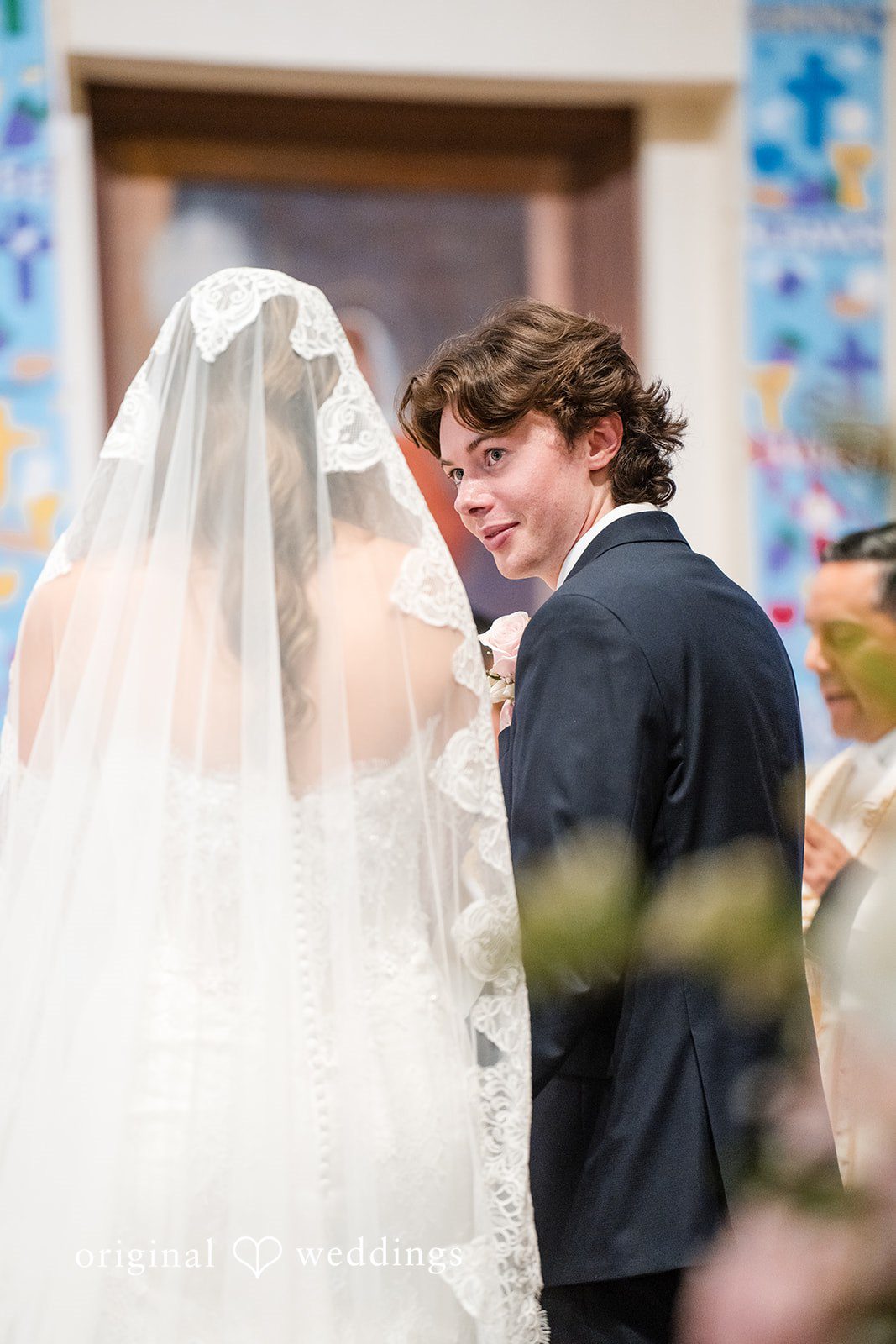 Maya + Max A groom smiling at his bride under her long veil as they face each other during the wedding service.