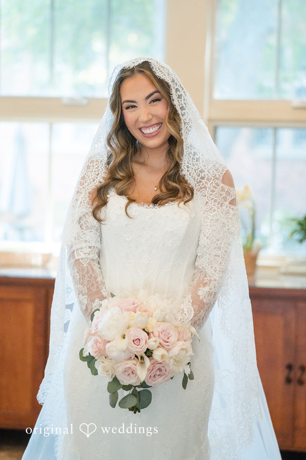 Maya + Max A smiling bride wearing a long lace veil and a white wedding dress, holding a bouquet of light pink roses.
