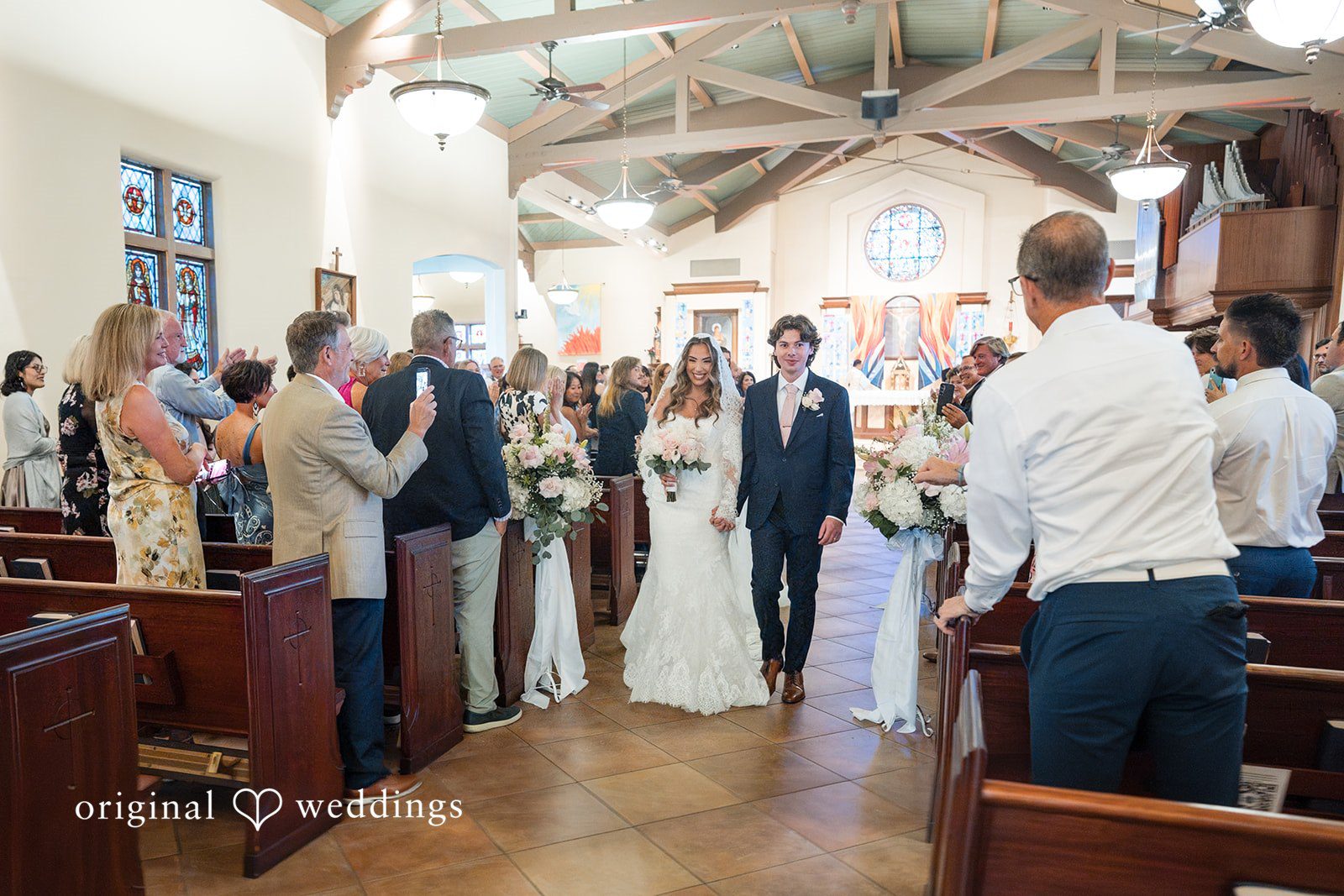 Maya + Max A bride and groom walking hand-in-hand down the church aisle together as guests clap and watch them exit.