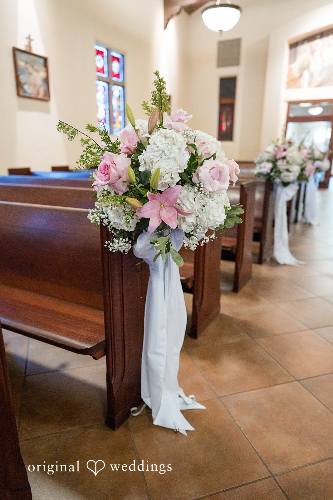 Maya + Max A close-up of a wooden church pew decorated with a large bouquet of pink and white flowers tied with a white fabric bow.