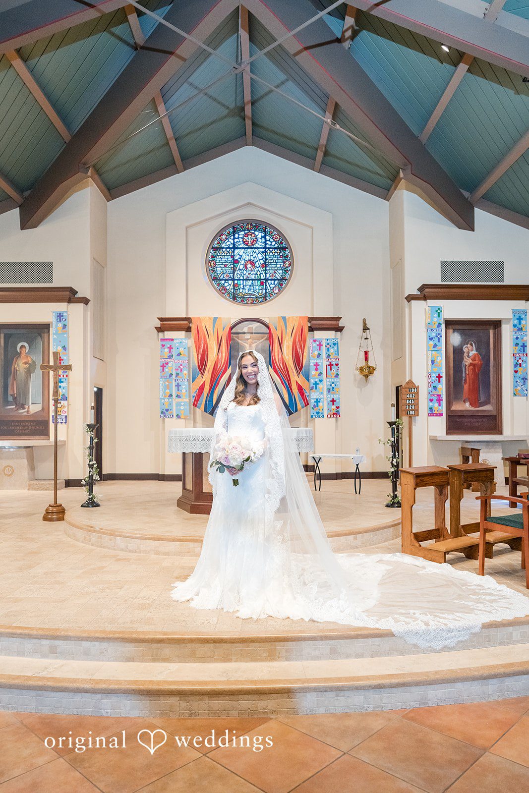 Maya + Max A bride standing alone inside a large church facing away from the camera toward the decorated altar.