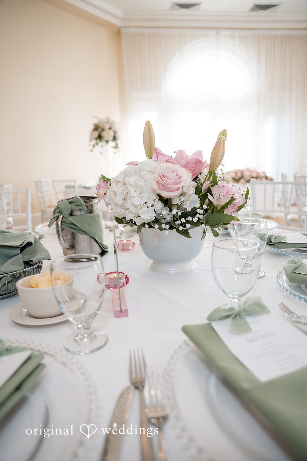 Maya + Max A close-up of a wedding reception table setting with a white tablecloth, green napkins, silverware, and a centerpiece arrangement of white and pink flowers.