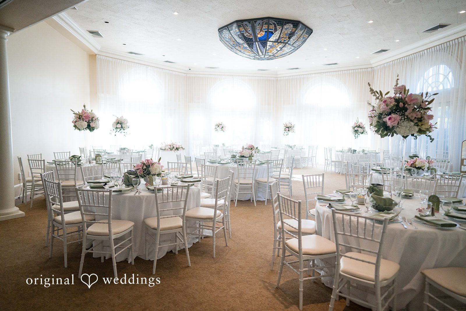 Maya + Max A wedding reception hall featuring round tables with white tablecloths, chairs, and elegant floral centerpieces under bright lighting.