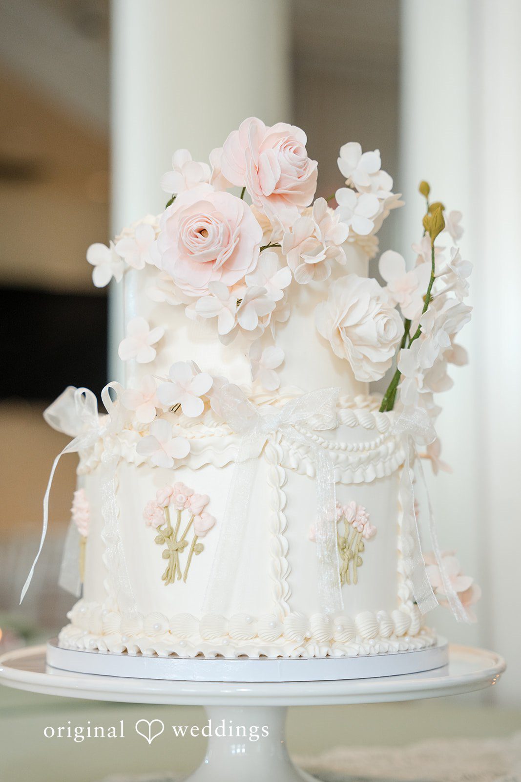 Maya + Max A multi-tiered white wedding cake decorated with delicate white frosting designs and topped with light pink flowers.
