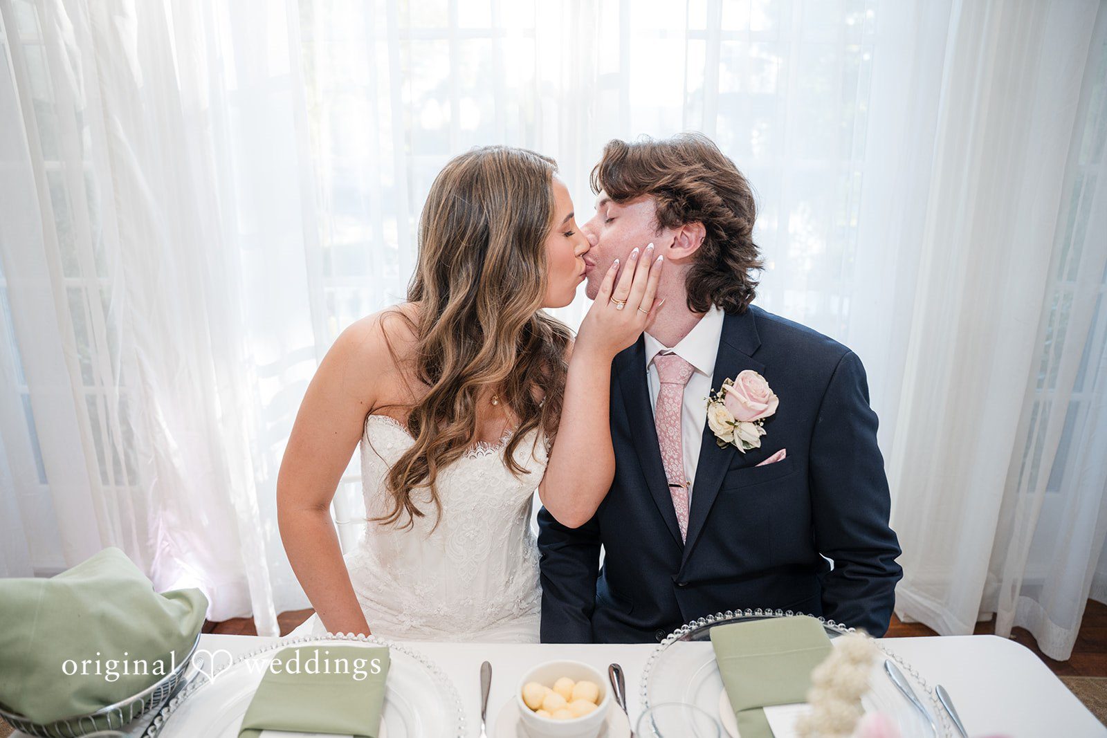 Maya + Max A bride and groom sharing a kiss while seated at their wedding reception head table.
